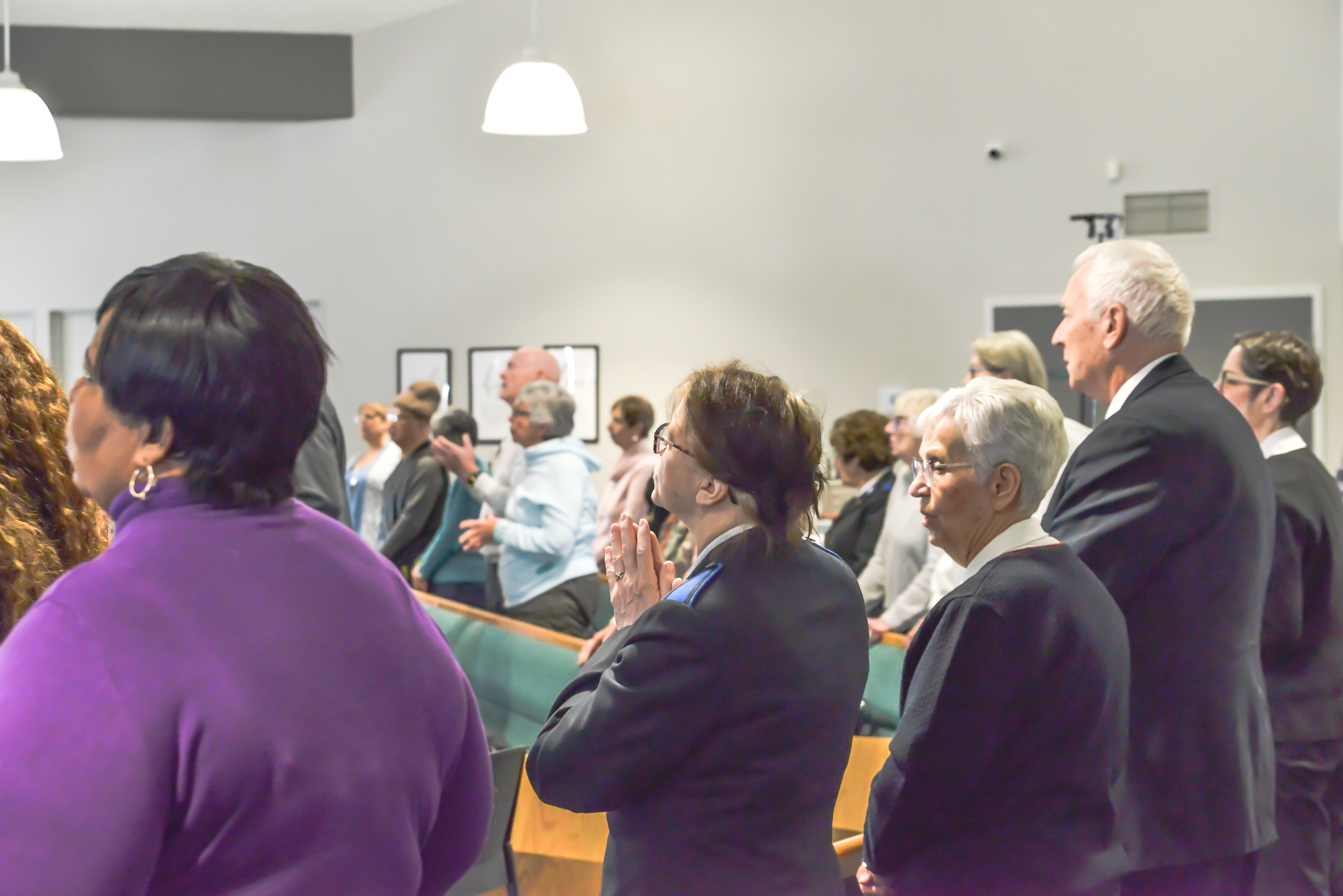 Congregation standing inside a church sanctuary facing the front, with rows of seating and people gathered closely together during a worship service.