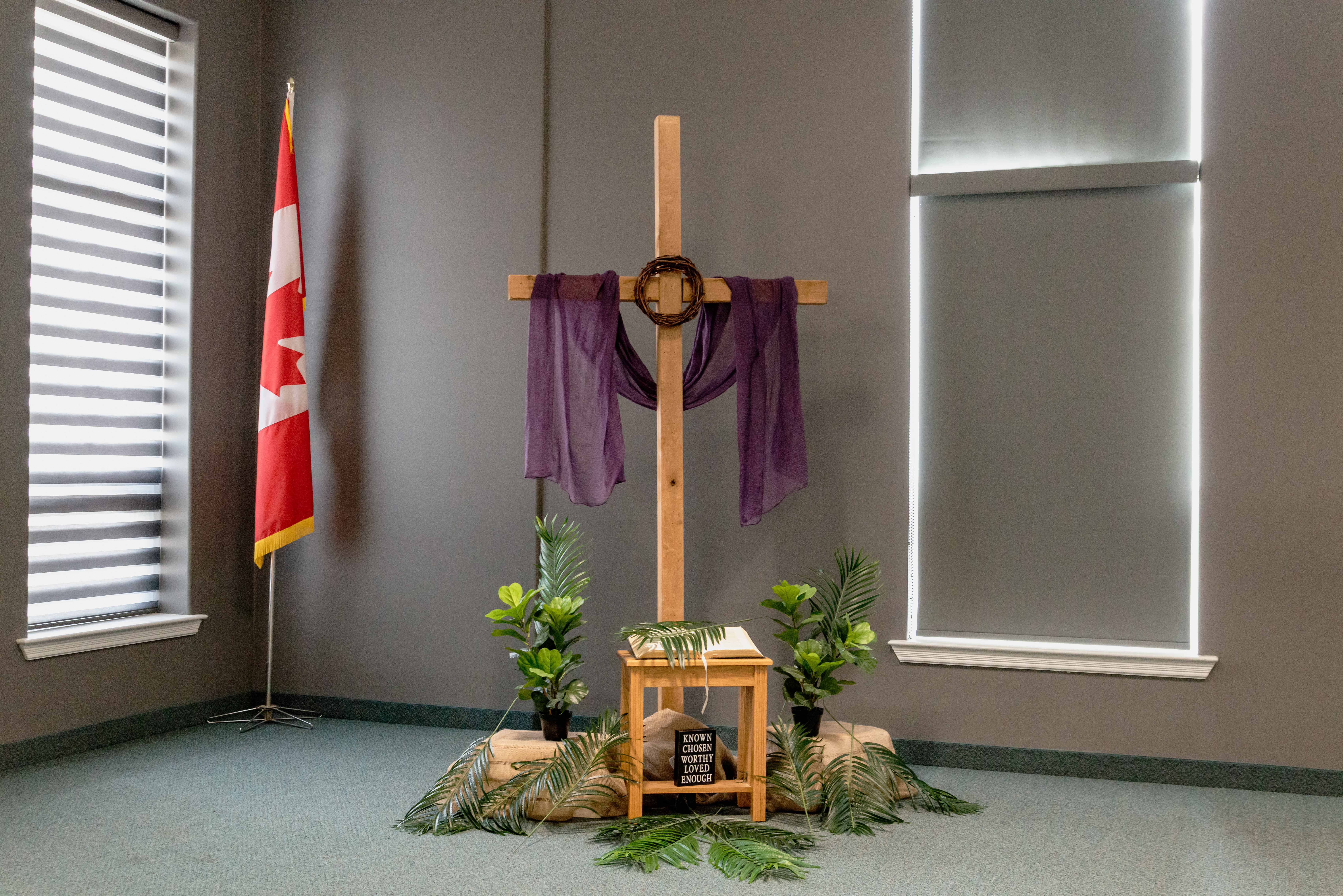 A wooden cross draped with purple cloth stands at the front of a church space, surrounded by potted plants and palm leaves, with a small table holding decorative items in front, and a Canadian flag positioned to the side near a window.